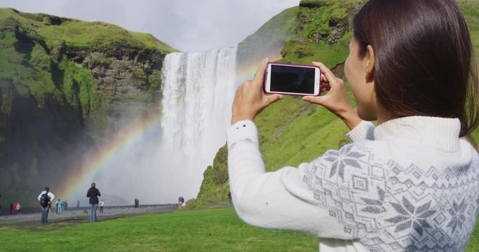 Woman tourist by waterfall Skogafoss on Iceland taking photo with smart phone outdoor. Girl visiting famous tourist attractions and landmarks in Icelandic nature landscape, ring road route 1.