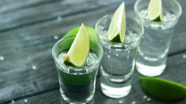Row of glass shots with tequila and green sour lime slices on wooden table with heap of salt