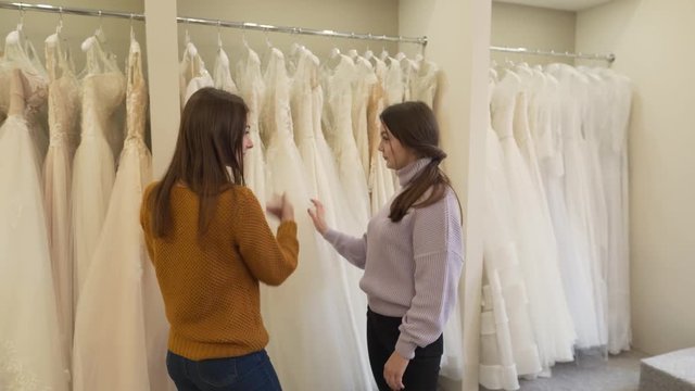 Two brunette girls are in wedding salon. One of them is future bride,another is her best friend. They are choosing dress for fitting.