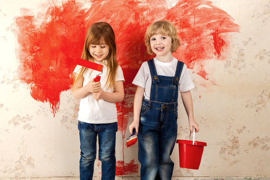 Children Painters. Small Artists With A Paint Brush In Their Hands And Paint Roller With Red Paint On The Wallpaper In The Room On The Wall.