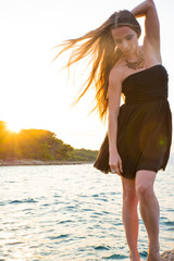 A beautiful woman in an elegant black dress standing and posing on the rocks next to the sea during the sunset.