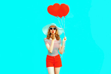 Beautiful young woman with red heart shaped balloons sending sweet air kiss on colorful blue background