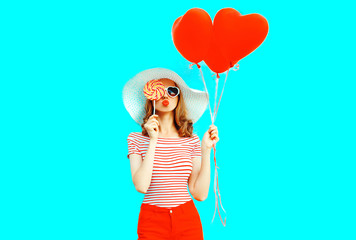 Beautiful young woman with red heart shaped balloons sending sweet air kiss closing her eye with lollipop on colorful blue background