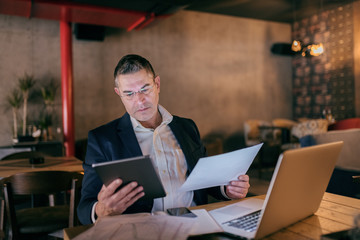 Caucasian businessman dressed smart casual holding tablet and paper and comparing data while sitting in cafe. On table laptop.