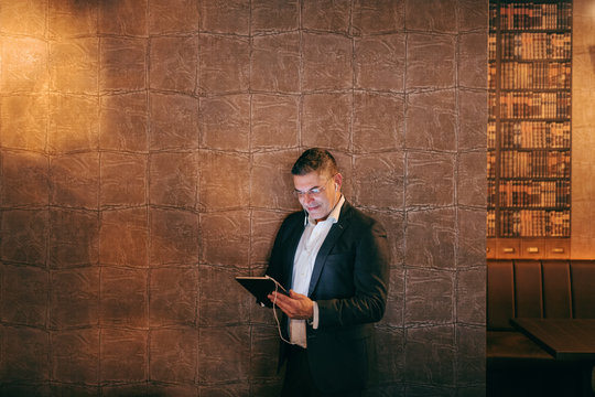 Middle-aged businessman standing indoors against the wall and using tablet for video call.