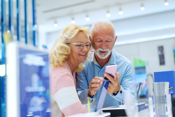 Close up of Caucasian old married couple trying out smart phone they want to buy while leaning on the stand. Tech store interior.