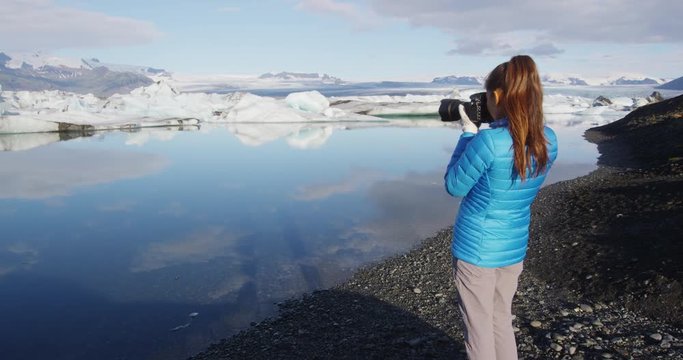 Photographer tourist woman taking photos with DSLR camera on travel on Iceland by Jokulsarlon glacial lagoon / glacier lake on Iceland. Happy tourist girl on travel in beautiful nature landscape.

