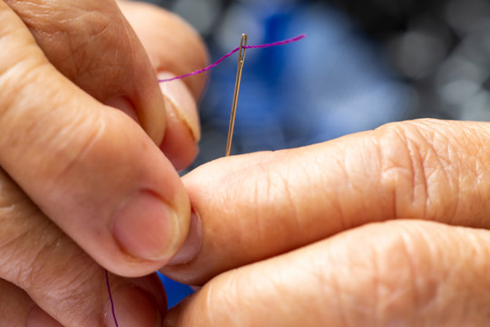 Senior woman's hands trying to thread a needle, Purple colour roll of thread, Close up & Macro shot, Selective focus, Blurred and bokeh background, Tailor, Needlework concept