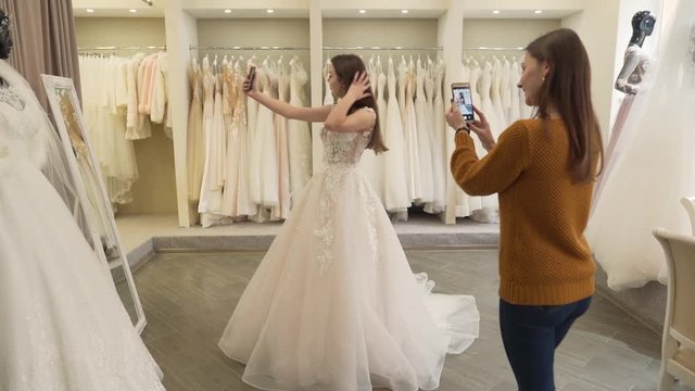 Two brunette girls are in wedding salon. They have a fitting of wedding dress. Both of them are holding phones in hands and making photos.