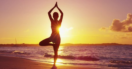 Yoga woman training in sunset in tree pose meditating outdoors on beach ocean sea. Female yoga instructor working out training in serene ocean landscape. Silhouette of woman model against sun. - Powered by Adobe