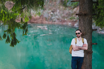 Handsome man over green nature and blue lake standing near the tree. Adventure, travel, tourism and leisure concept - young boy relaxing in woods.