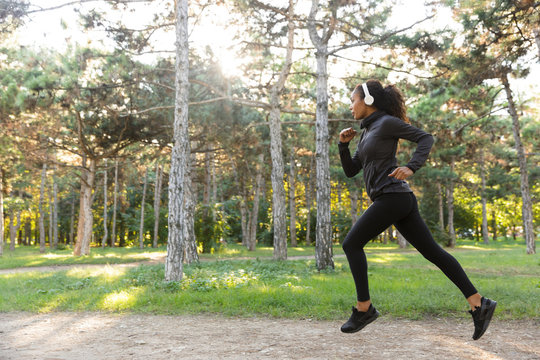 Photo of sportswoman 20s wearing black tracksuit and headphones working out, while running through green park