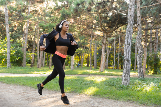 Image of american woman 20s wearing black tracksuit and headphones working out, while running through green park