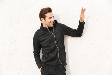 Image of handsome sportsman 30s in black sportswear and earphones, standing over white wall outdoor