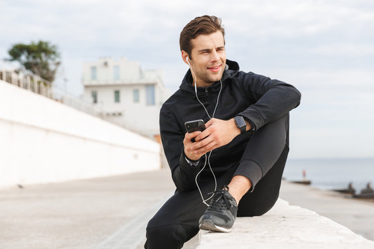 Image of european sportsman 30s in black sportswear, using earphones and mobile phone while sitting on boardwalk at seaside