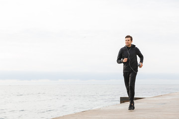 Image of sporty man 30s in black sportswear and earphones, working out and running by seaside