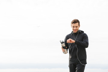 Image of satisfied sportsman 30s in black sportswear and earphones, working out and running by seaside