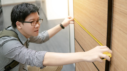 Young Asian man using tape measure for measuring modern wooden cabinet in walk-in closet showroom....