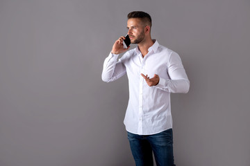 A serious handsome young man in a white shirt talking on his phone and standing in front of a grey...