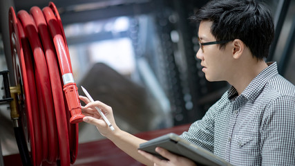 Young Asian male technician checking red fire hose reel by using digital tablet and pen. Building...