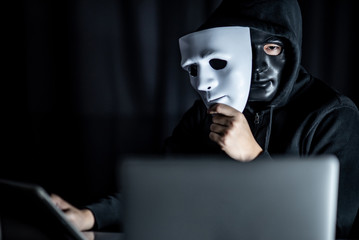 Mystery male hoodie hacker wearing black mask holding white mask sitting with laptop computer on...