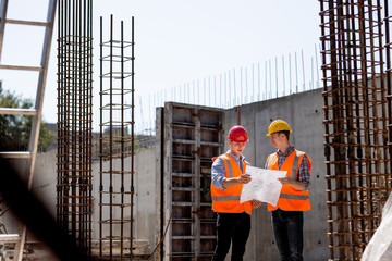 Civil engineer and construction manager in orange work vests and hard helmets explore construction documentation on the building site near the steel frames