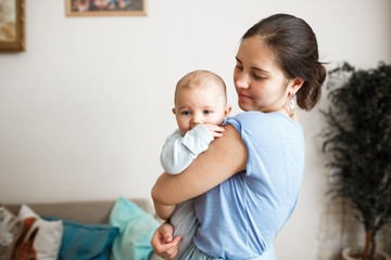 Young mother dressed in light blue t-shirt and skirt is holding her tiny son on her arms in the room at home