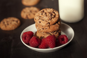 Chocolate chip cookies on white plate dark old wooden table with red raspberry and milk. Delicious breakfast.