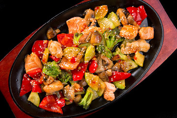 Fish salad with salmon pieces, vegetables, mushrooms and sesame, in a pan, on a black background