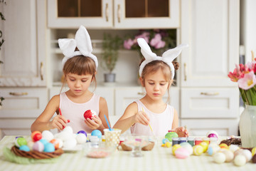 Obraz na płótnie Canvas Two little sisters with white rabbit's ears on their heads dye the eggs for the Easter table in the cozy light kitchen