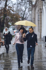 Naklejka premium Beautiful couple, guy and his girlfriend dressed in casual clothes are running under the umbrella on the street in the rain.
