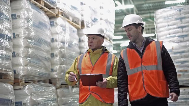 Workers walking and discussing stock inventory against tall shelves, in a warehouse with bright lamps.