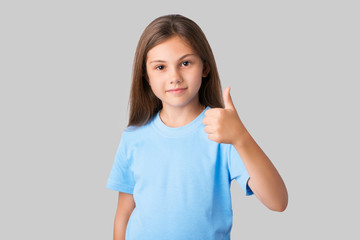 Young girl in blue t-shirt with long natural hair showing thumbs up against a grey background. Portrait of a smiling small schoolgirl