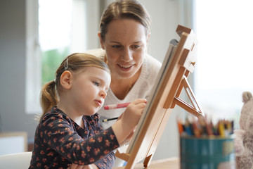 Mother and young daughter drawing together at home on easel