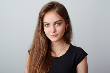 Close-up shot of beautiful smiling young female poses in studio against a grey background. Has natural long hair, grey eyes and pure skin. Wears black shirt, gold chain and earrings.