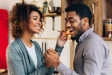 Woman in kitchen letting man taste meal with wooden spoon