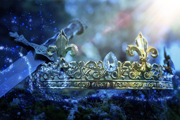 mysterious and magical photo of silver king crown and sword over the stone covered with moss in the...