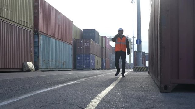 Caucasian man in white hardhat talking by cellphone walking through dockyard containers.