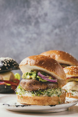 Variety of homemade classic, vegan and mini burgers in wheat and black buns with beef and veal cutlets, portobello mushroom, avocado, melted cheese and vegetables on white marble table.