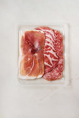 Antipasto meat platter assorti of sliced jamon, salami sausage in opened plastic packaging over white marble background. Flat lay, space