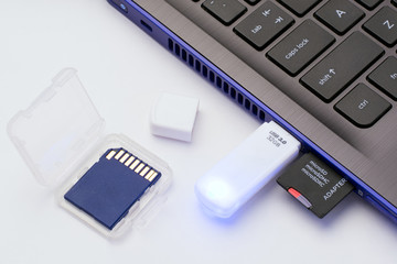 MicroSD memory card with adapter, white USB stick plugged in the ports of the modern blue laptop. Blue led on the USB stick is on. Blue SD memory card in a transparent case lays near the computer.