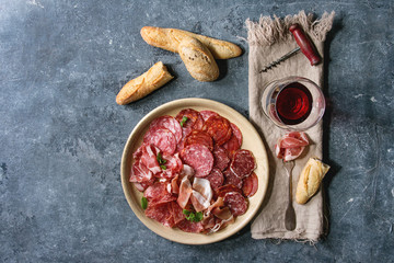 Antipasto meat platter assorti of sliced jamon, salami, chorizo sausage in ceramic plate with bread and glass of red wine on cloth over blue texture background. Flat lay, space - obrazy, fototapety, plakaty