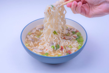 Curvy Ramen noodles being pulled up by chopsticks. Asian Korean soup with mushrooms and vegetables in blue bowl on white background.