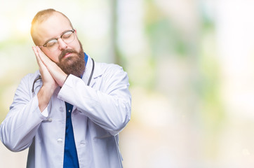 Young caucasian doctor man wearing medical white coat over isolated background sleeping tired dreaming and posing with hands together while smiling with closed eyes.