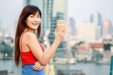 Rear view of a smiling young woman standing at bangkok skyline with a cup of coffee after working in the evening..