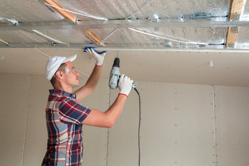 Young man in goggles fixing drywall suspended ceiling to metal frame using electrical screwdriver...