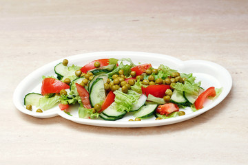 Original salad for healthy nutrition of cucumbers, tomatoes, lettuce and green peas on a white plate with olive oil, sprinkled with pepper on a marble table