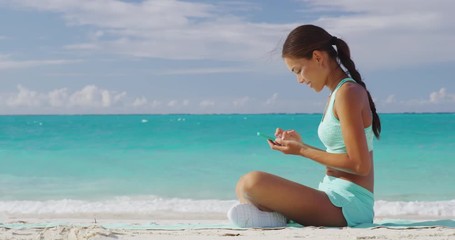 Fit girl using fitness app on phone during travel holidays on beach texting or posting on social media online. Healthy sporty Asian woman living a happy active life exercising on towel. - Powered by Adobe
