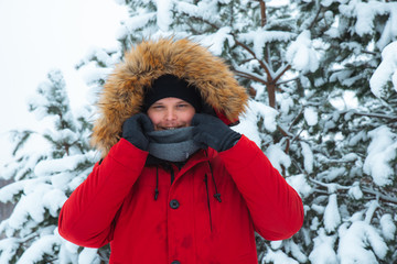 young adult man in red winter coat with hood with fur. snowed fir-tree on background