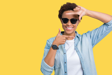 Fototapeta na wymiar Beautiful young african american woman wearing sunglasses over isolated background smiling making frame with hands and fingers with happy face. Creativity and photography concept.